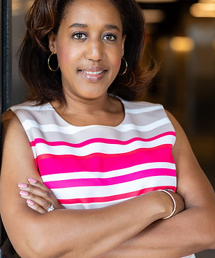 A picture of the founder of the company. She is a black woman with shoulder length hair, wearing a striped pink and white dress. She is looking into the camera, smiling, with her arms folded.