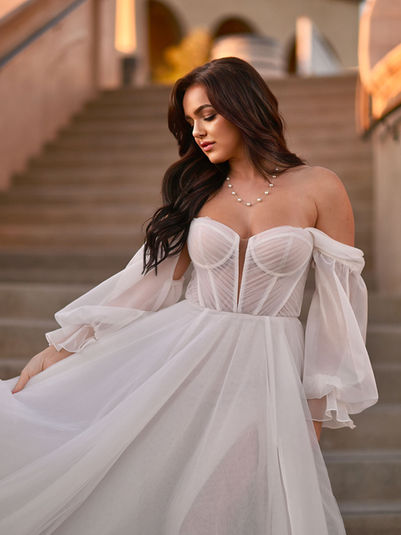 1497 by Martina Liana
LUXE STRAPLESS A-LINE WEDDING DRESS WITH OFF-THE-SHOULDER BELL SLEEVES
Turn heads in this soft luxe bridal look. Style 1497 is the perfect blend of modern luxury with the contoured bodice of exposed seaming and the delicate beauty of the voluminous layered skirt. The pleated semi-sheer strapless bodice offers an illusion plunging neckline with flattering angles that taper the waist. The detachable off-the-shoulder sleeves gather at the wrist for a romantic couture allure. In the back bodice, the sheer pleats resolve into a row of fabric-covered buttons before meeting the gathered soft A-line skirt. Finally, a high side slit emanates a luxurious romanticism for the fashion-forward bride.