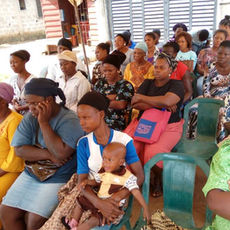 African women studying in an open-air classroom. A woman in front holds a baby.