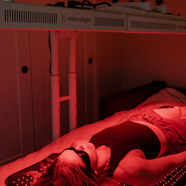 A woman lies underneath a red light panel and lies on top of a red light therapy mat.