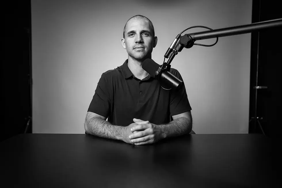 Dr. Andy Galpin in a black shirt sits at a table with a microphone, posing confidently against a plain backdrop. Black and white photo.