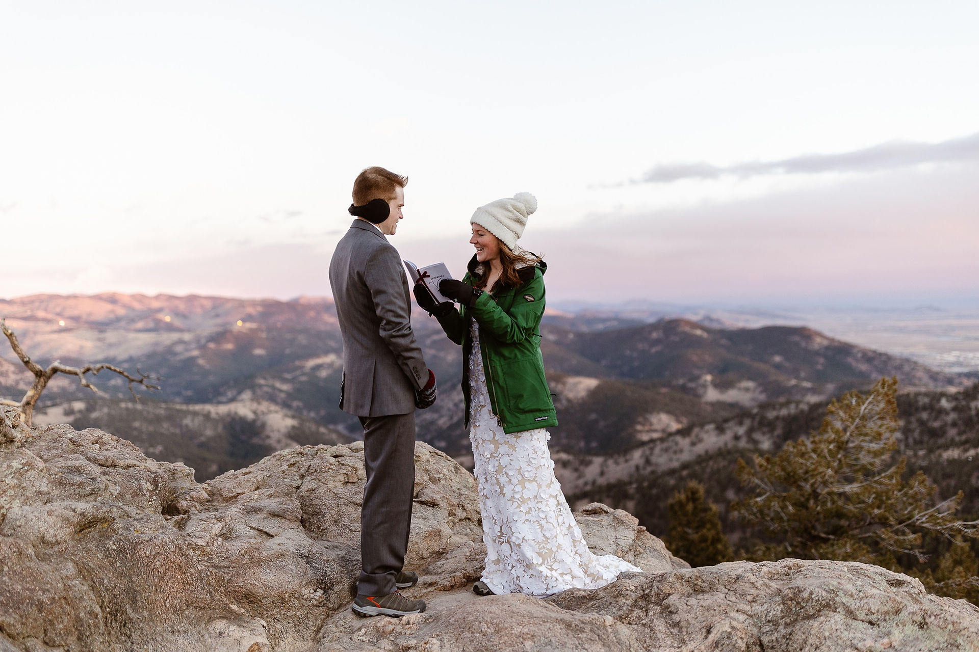 A couple stands together on a rocky overlook during an outdoor ceremony, surrounded by mountain views and soft evening light.