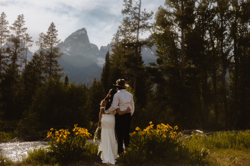 A couple embracing with backs turned to camera in a riverfront bank of yellow flowers, looking toward forest and mountain scenery.