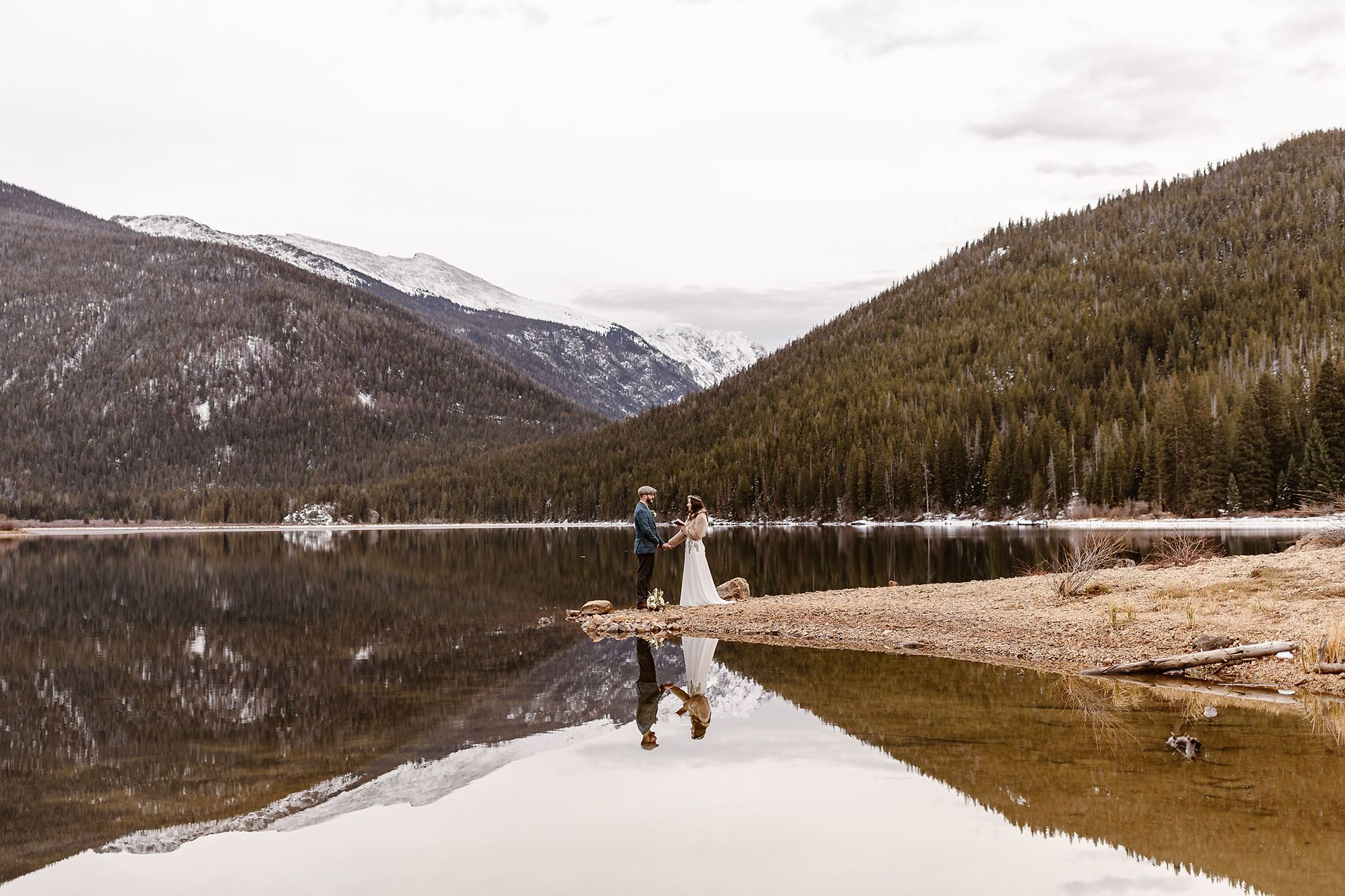 A couple stands together on a narrow lakeshore with snow-covered mountains and evergreen forests reflected in the still water around them.