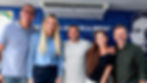 Cinco pessoas sorrindo posam em frente a um banner azul com texto parcial em português, em clima descontraído.