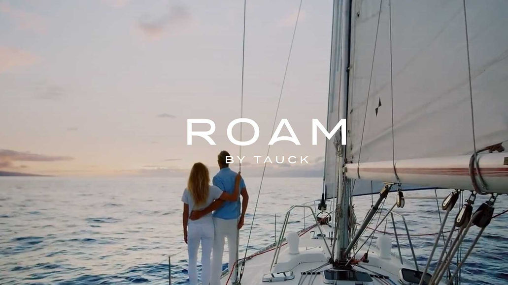Couple standing arm in arm on a sailboat deck at sunset with the word ROAM overlaid