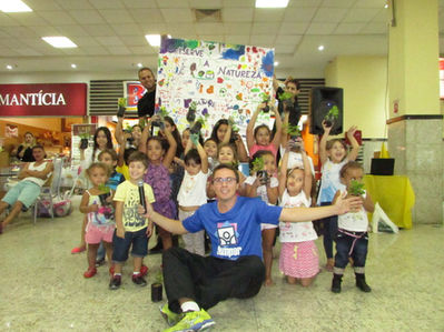 Group of children holding plants and smiling, with a banner EVENTO SOCIAL E FESTA INFANTIL.