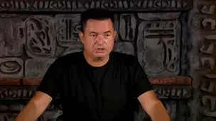 Adam siyah tişörtle, arka planda dekoratif taş duvar önünde duruyor. Ciddi bir yüz ifadesi var, ortam loş ve gizemli.