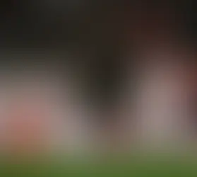 Futbolcular kırmızı-beyaz çizgili formalarla gol sevinci yaşıyor. Seyirciler arka planda. Sahada coşkulu bir atmosfer var.