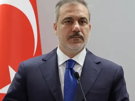 Önde mikrofonla takım elbiseli bir adam. Arkada Türk bayrağı. Resmi ortam, ciddi ifade. Arka plan sade ve açık renk.