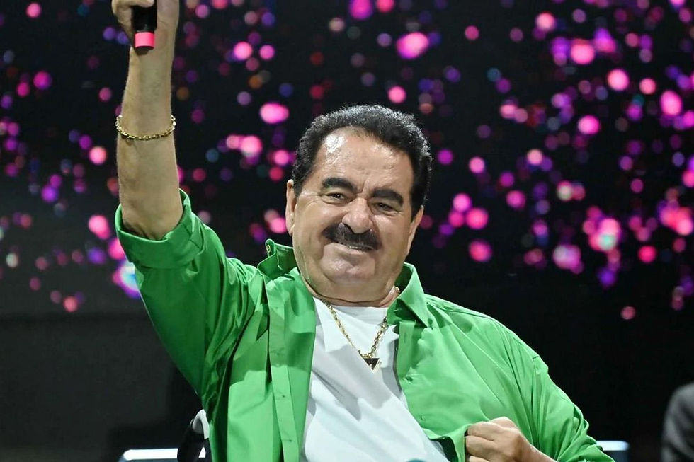 Yeşil gömlekli, mikrofonlu bir adam sahnede elini kaldırıyor; arkada pembe ışıklar ve neşeli bir atmosfer var.