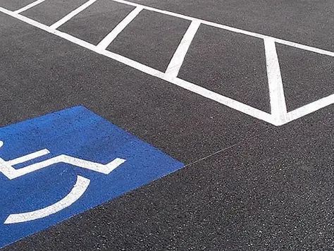 Mavi zeminde beyaz tekerlekli sandalye simgesi ve yanındaki eğik beyaz çizgiler, siyah bir otopark alanında dikkat çekiyor.