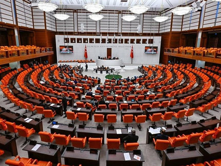 Türkiye Büyük Millet Meclisi genel kurul salonu; turuncu koltuklar daire şeklinde dizilmiş, üyeler çalışmalar yapıyor. Arka planda Türk bayrağı.