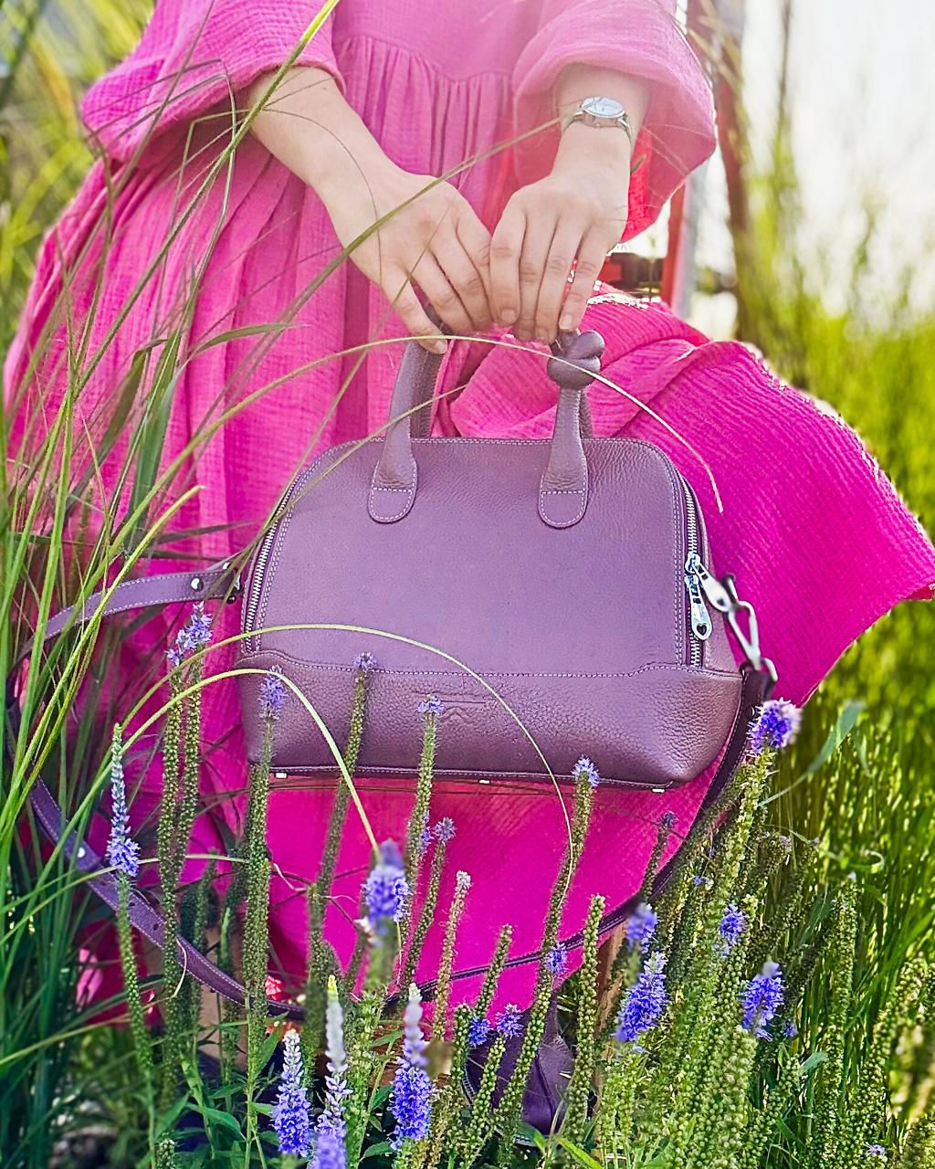 Жіноча шкіряна сумка рожевого кольору з короткими ручками у відтінку аметист на фоні літнього пейзажу
