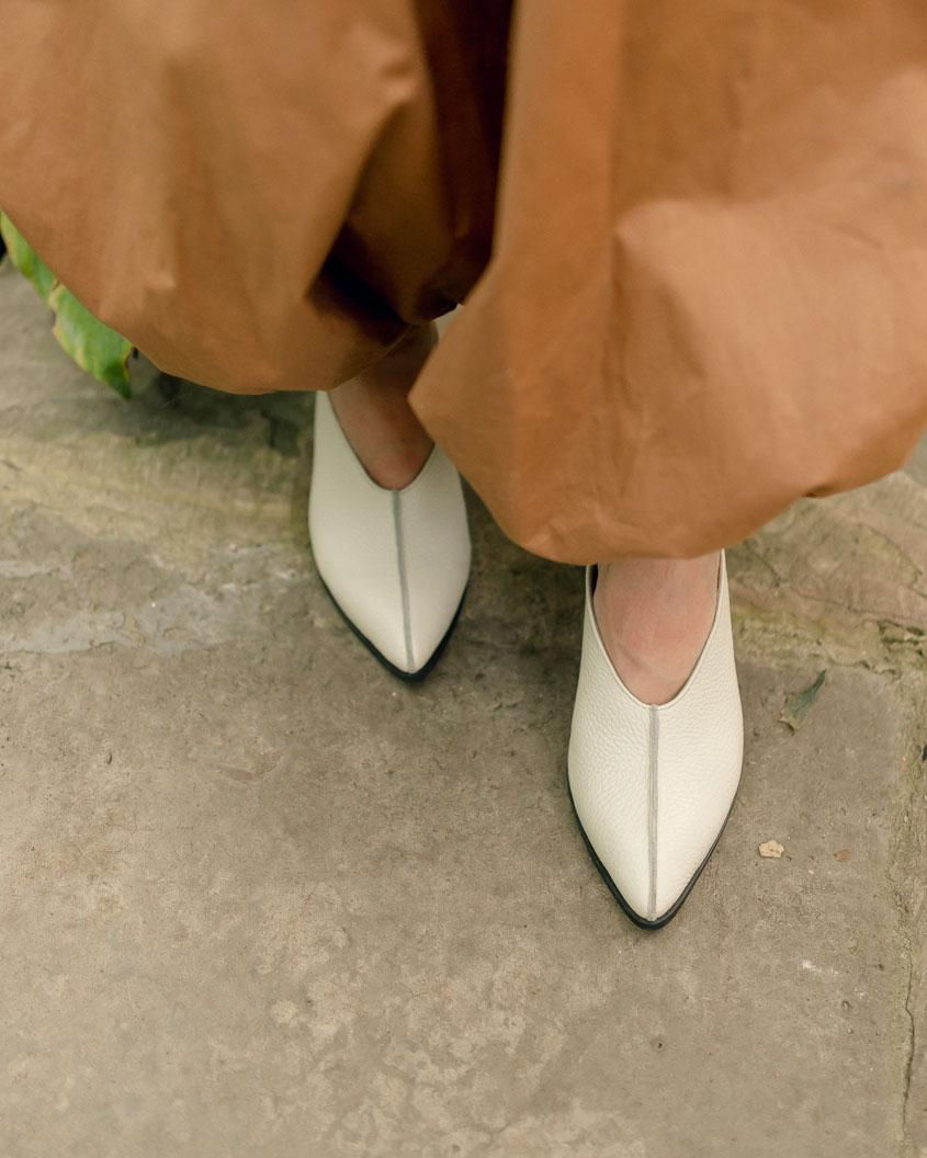 Прев'ю: Стильні жіночі сабо молочного кольору з гострим носком та центральним швом на ногах моделі на кам'яній підлозі.