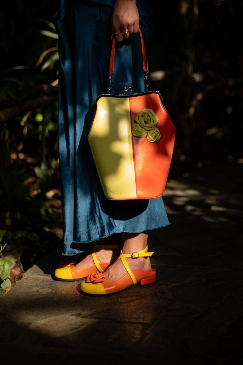 Thumbnail: Kobieta w granatowej spódnicy prezentuje kolorowe sabo skórzane z kwiatem na kamiennej ścieżce.