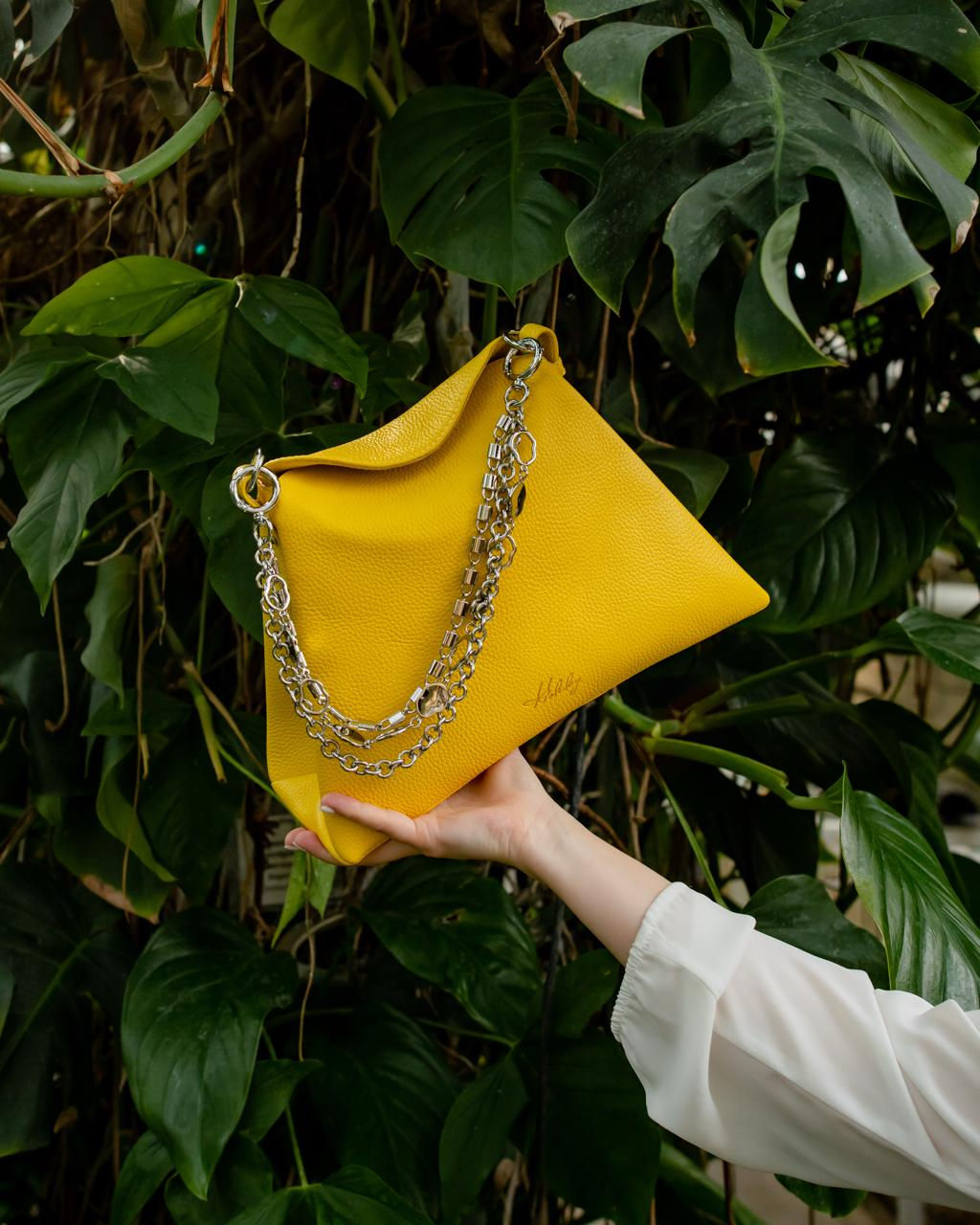 Жіноча шкіряна сумка яскраво-жовтого кольору з багатошаровим декоративним ланцюжком на фоні тропічних рослин.