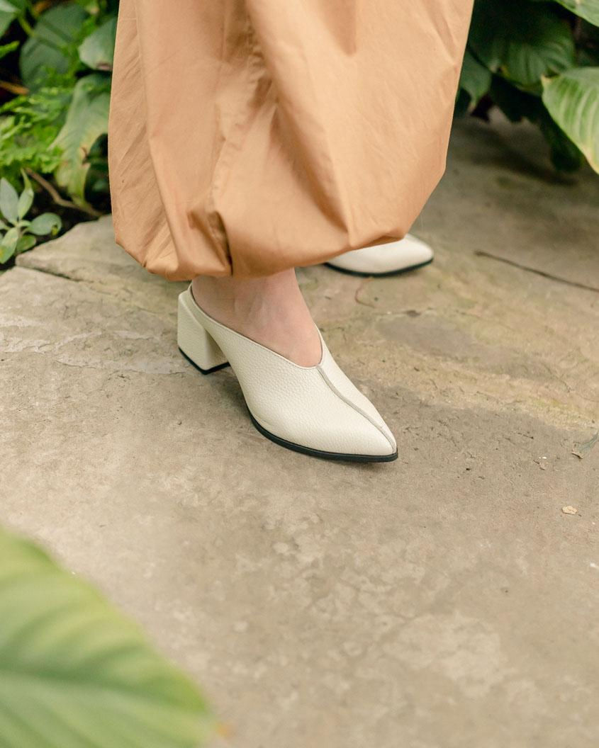Стильні жіночі сабо молочного кольору з гострим носком та центральним швом на ногах моделі на кам'яній підлозі.