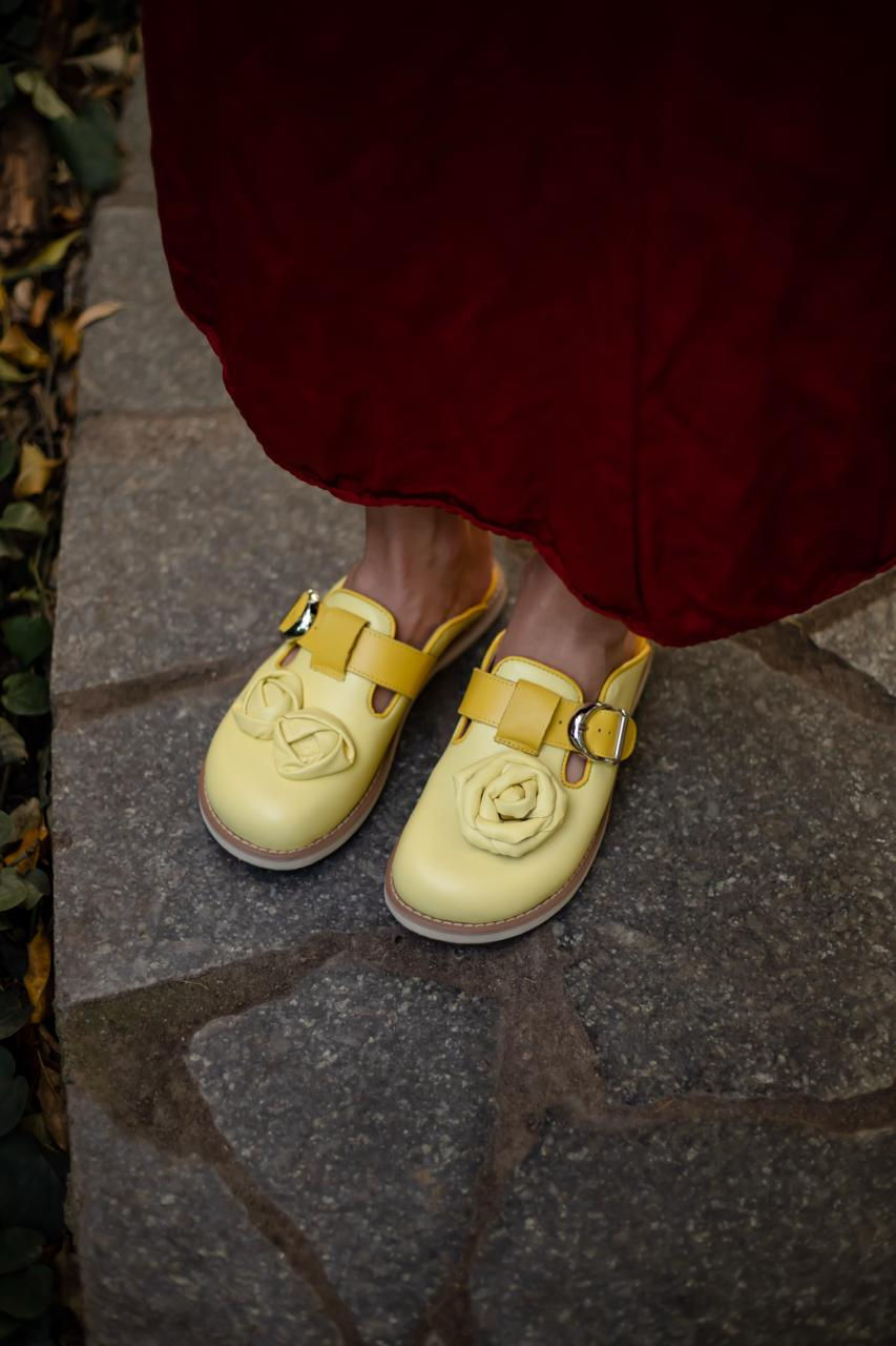 Kobieta prezentuje skórzane chodaki z trójwymiarową różą na kamiennej ścieżce, widok na artystyczny detal.