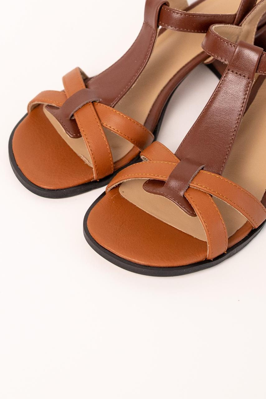 Thumbnail: Kobieta prezentuje brązowe skórzane sandały T-strap na niskim obcasie na tle kamiennej ścieżki.