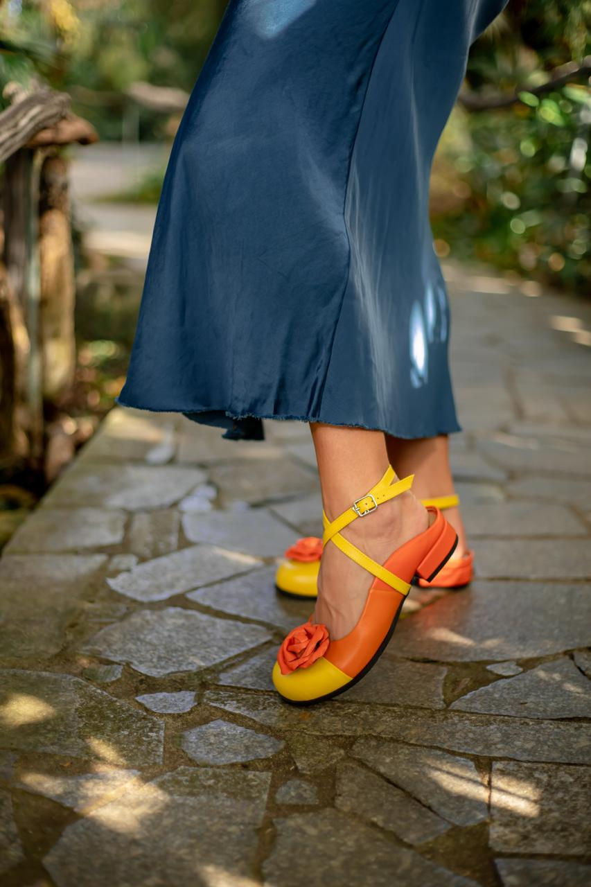 Thumbnail: Kobieta w granatowej spódnicy prezentuje kolorowe sabo skórzane z kwiatem na kamiennej ścieżce.