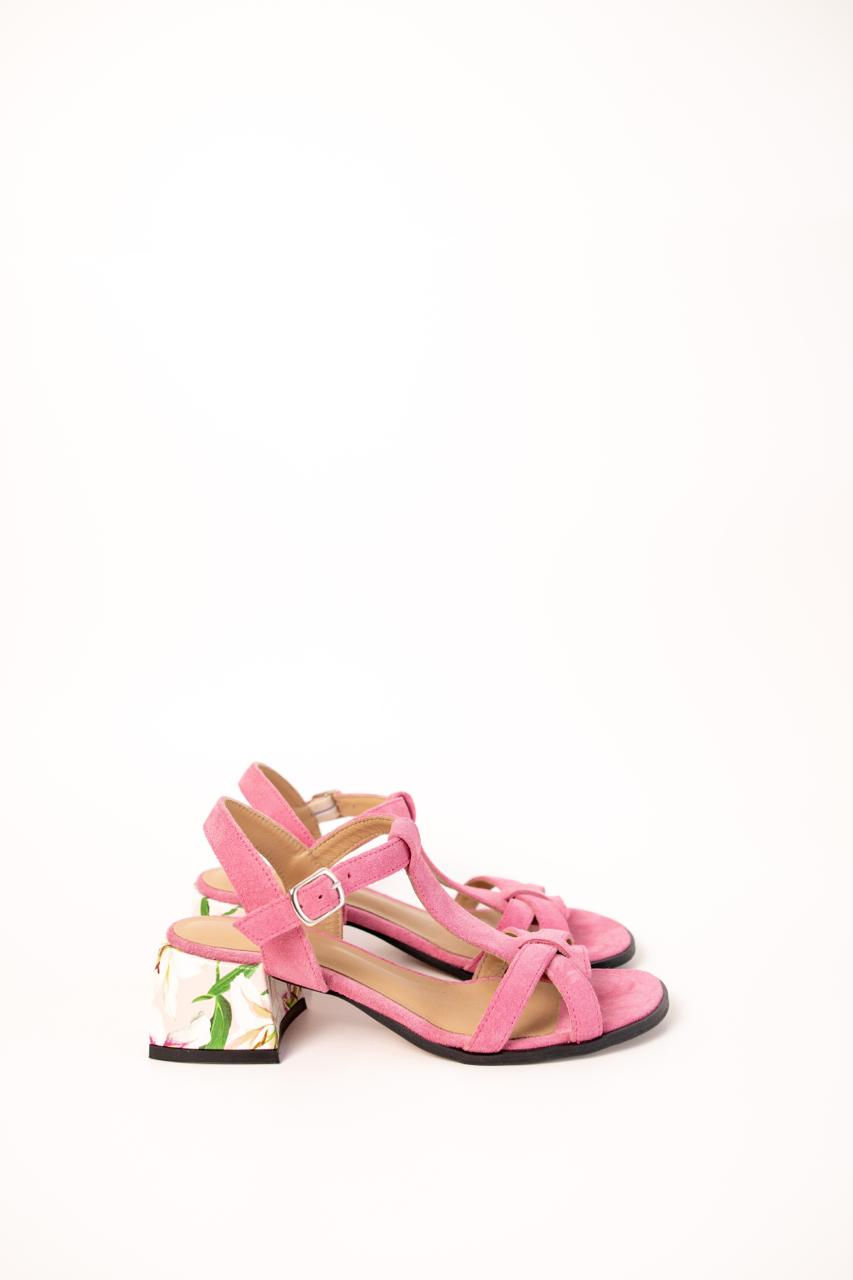 Kobieta w kwiecistej spódnicy prezentuje różowe zamszowe sandały na wzorzystym obcasie w otoczeniu zieleni.