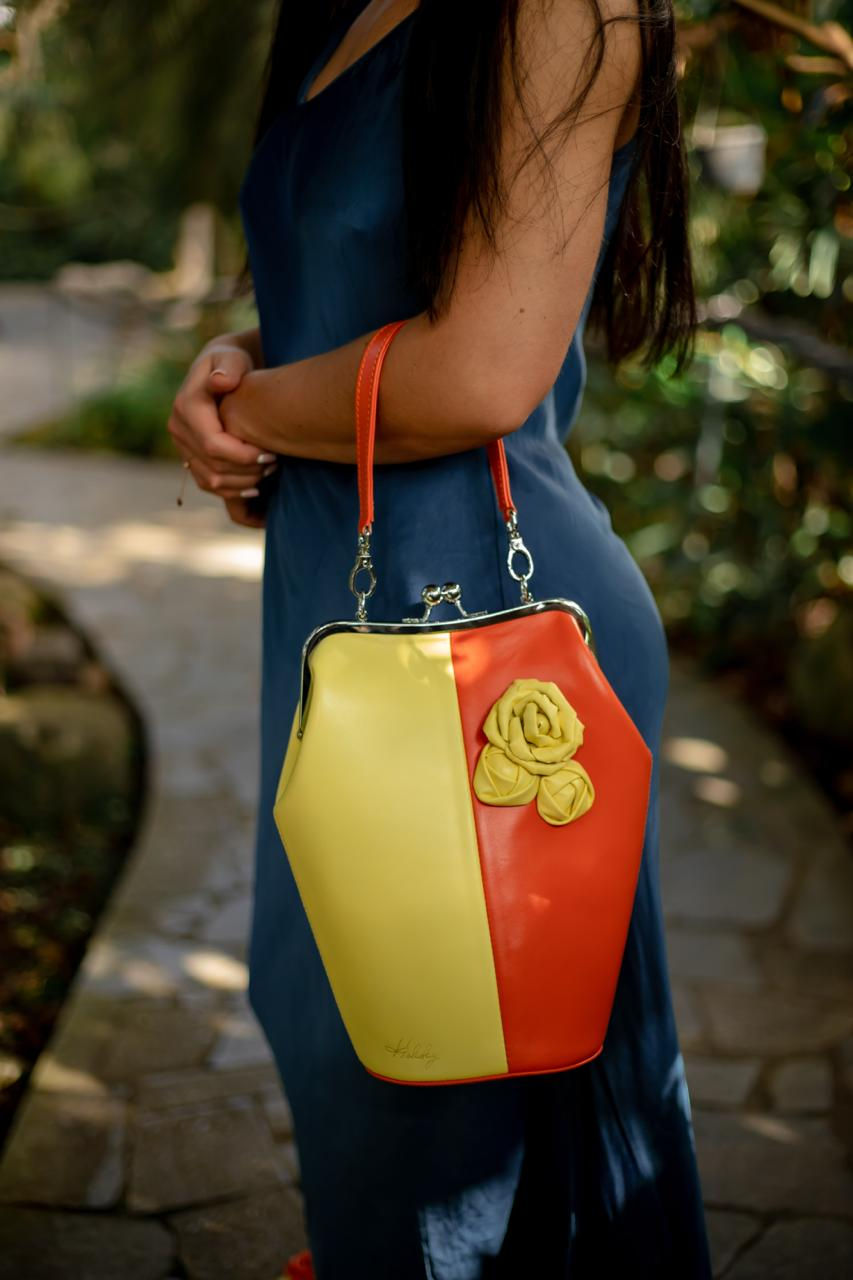 Miniatura: Dwukolorowa żółto-pomarańczowa torebka skórzana z ozdobną żółtą skórzaną różą i metalowym zapięciem, trzymana oburącz na tle