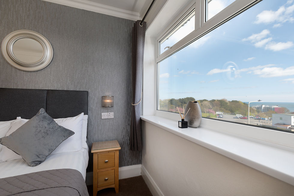 Modern bedroom with gray accent wall, circular mirror, and window view of trees and sky. Vase and diffuser on window sill, gray pillow on bed.