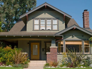 Craftsman-style house with gray siding, brick chimney, and porch. Surrounded by greenery and shrubs under a clear blue sky.