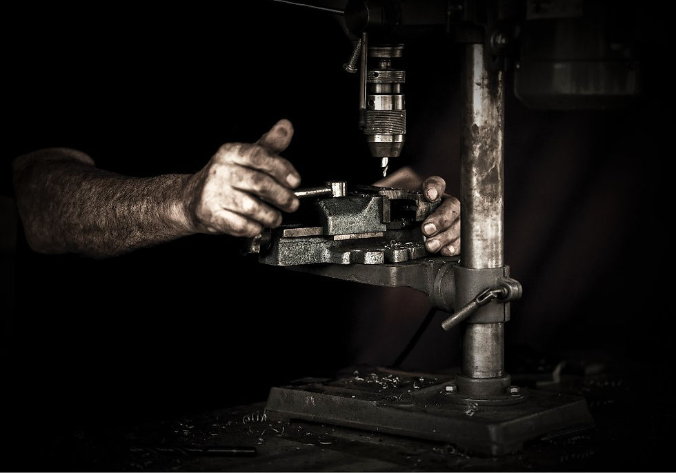 Hands operating a drill press in a dimly lit workshop. The worker's hands are greasy, focusing on a metal piece. Industrial mood.