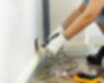 Person in gloves using a multimeter to test a wall socket in a room. Tools and electrical components scattered.