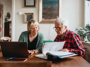Couple de seniors consultant des documents et un ordinateur à domicile, illustrant l’accompagnement administratif et le quotidien des retraités.
