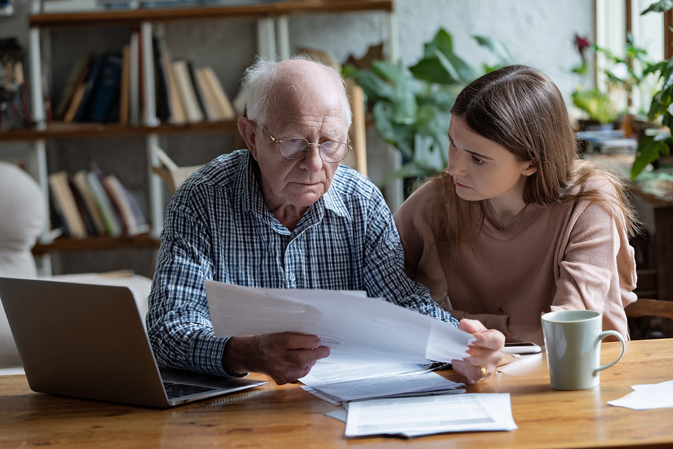 Un homme âgé concentré lit des documents aux côtés d’une jeune femme, dans un cadre familial, avec un ordinateur portable sur la table.