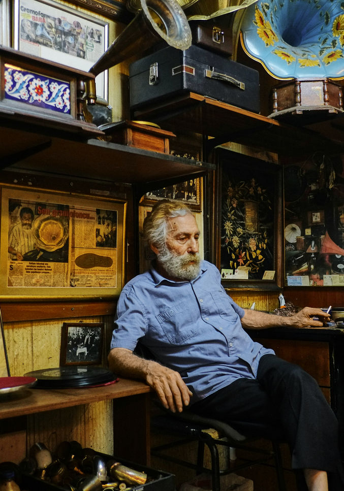 Elderly man with beard sitting in cluttered workshop surrounded by antique posters and objects.