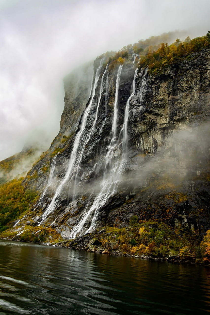 Wasserfall "Die sieben Schwestern" stürzt von einer nebligen Felsklippe in den Geirangerfjord; herbstliche Bäume oben.