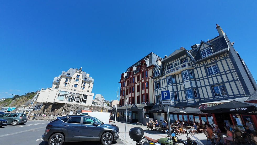 Straßenansicht mit Autos, Fachwerkhäusern und Café mit Besuchern unter Sonnenschirmen. Blauer Himmel, entspanntes Sommerflair. Granville am Casino