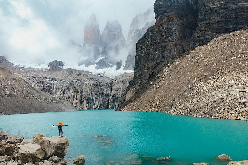 Eine Person in oranger Jacke steht auf einem Felsen am türkisfarbenen See vor nebligen, schneebedeckten Bergen in rauer Landschaft. Reiseziel Patagonien