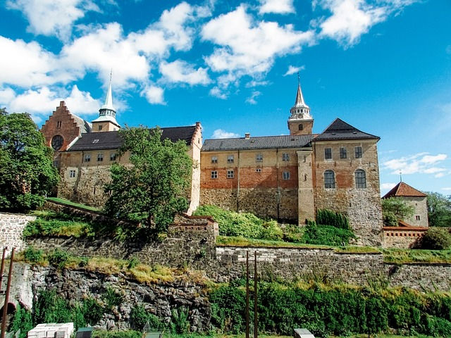 Historische Burganlage auf einem Hügel mit Türmen in Oslo, Steinmauern und grüner Böschung unter blauem Himmel.