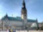 Historisches Rathaus mit hohem Uhrturm und blauem Dach; Passanten auf dem Platz, rechts ein 20er-Schild.