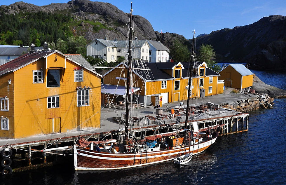 Gelbe Holzhäuser und ein Segelboot am Kai auf den Lofoten in einer norwegischen Fjordbucht, dahinter Berge und blauer Himmel.