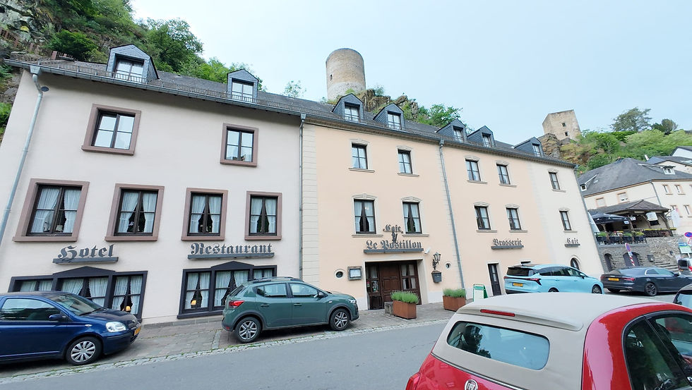 Hotel, Restaurant, Café Le Rostillon an Hangstraße mit geparkten Autos und Ruinentürmen im Grünen.