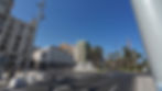 Sonniger Stadtplatz mit Springbrunnen, Palmen, spanischer Flagge und historischen Häusern unter blauem Himmel.