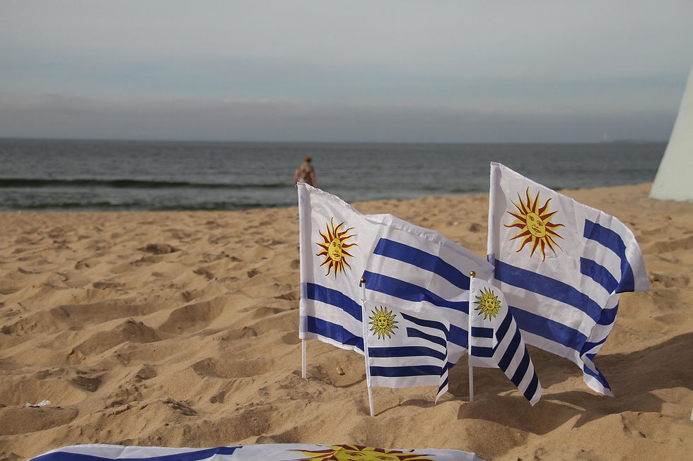 Uruguay-Flaggen im Sand am Strand, dahinter das Meer. Eine Person geht in der Ferne, friedliche Atmosphäre.