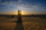 Stein in Wüste bei Sonnenuntergang, Sonnenstrahlen leuchten durch Felsöffnung. Landschaft in gelb und blau, Wolken am Himmel.