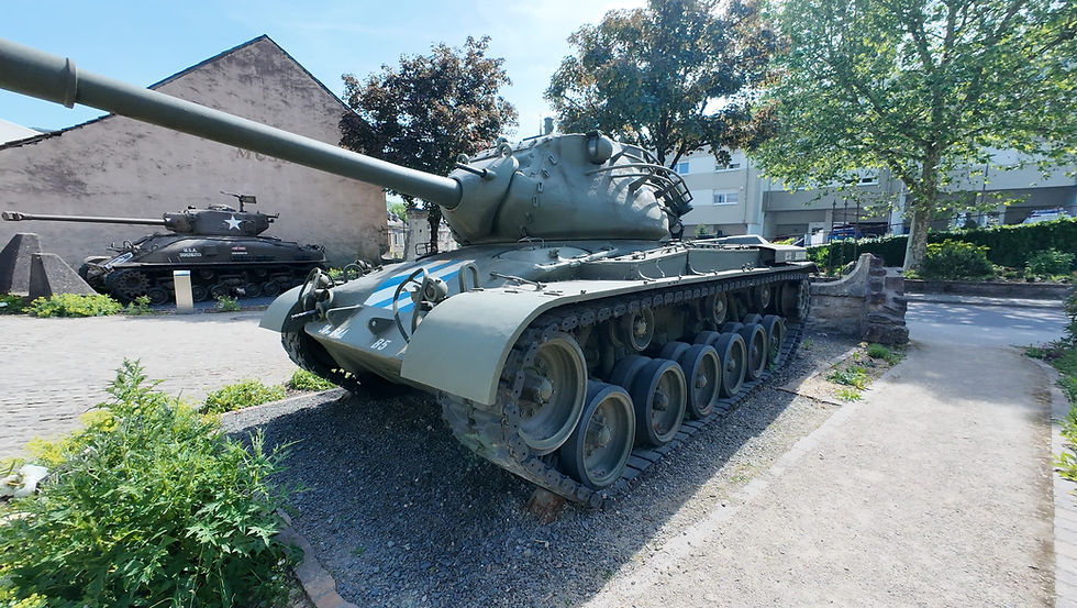 Two tanks on display at a sunny museum: a large green tank in front and a black U.S.A. tank behind, with trees and buildings.