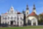 Historische Kirche und prunkvolles Gebäude mit Türmen unter blauem Himmel; drei Menschen gehen davor über den Platz.