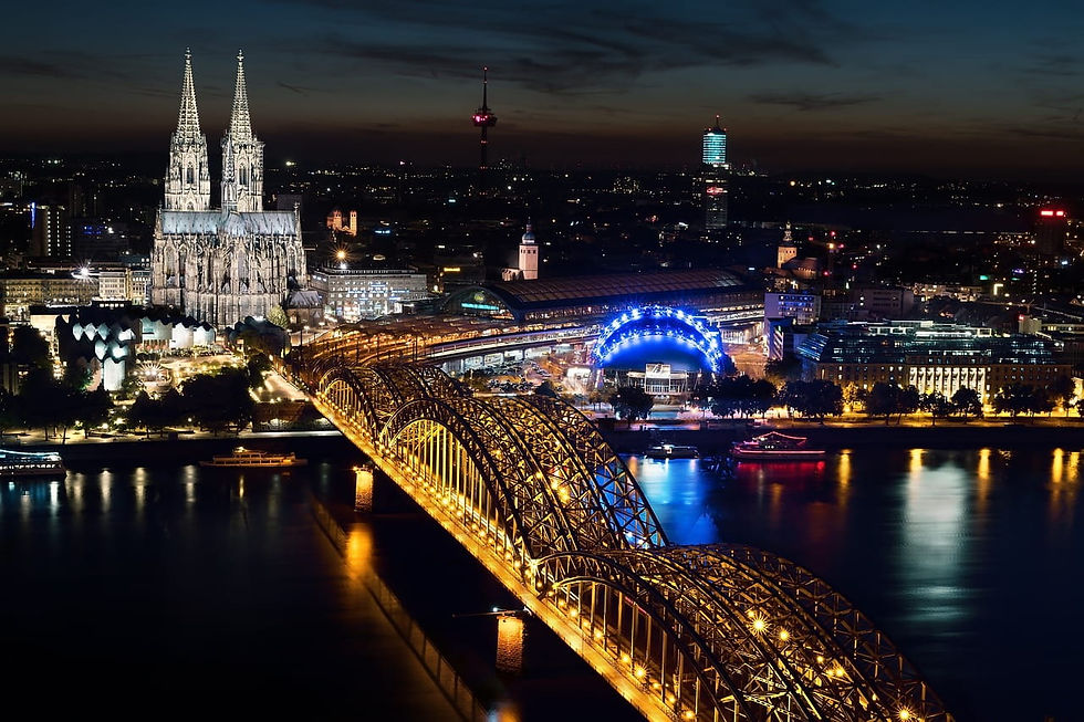 Nächtliche Stadtansicht mit beleuchtetem Dom, goldener Brücke und blauem Rundbau. Reflektionen im Wasser erzeugen eine stimmungsvolle Atmosphäre.