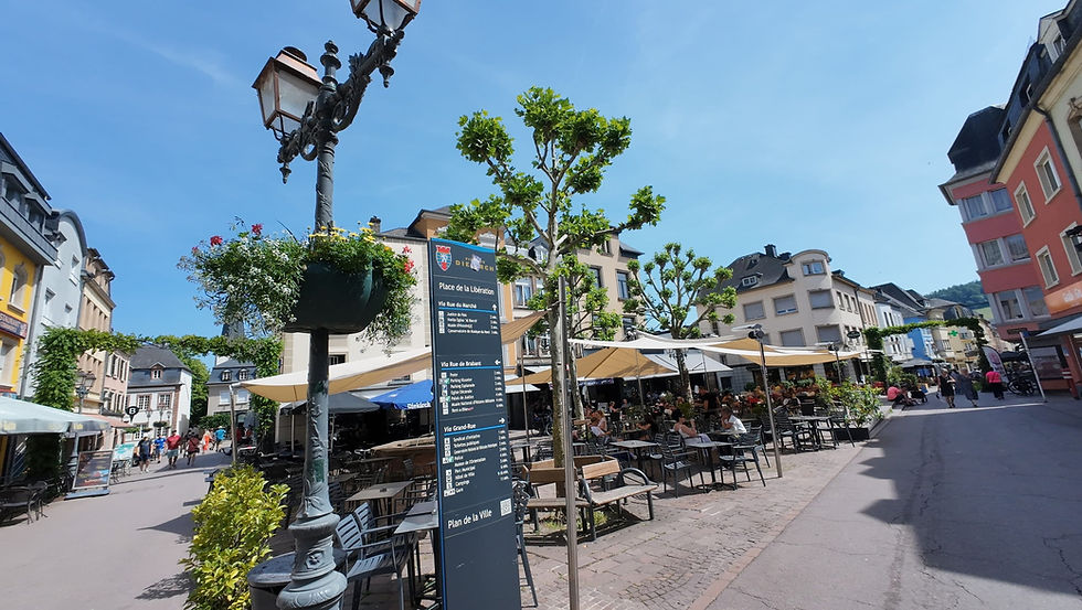 Sunny European plaza with café tables, planters, and a sign reading Place de la Libération in Diekirch; people relax outside.