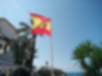 Spanische Flagge weht an Mast zwischen Palmen über Küste und Meer unter wolkenlosem Himmel.