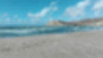 Sandstrand mit sanften Wellen, klarem blauem Himmel, weißen Wolken und einem Hügel im Hintergrund. Einige Fußspuren im Sand. Ruhige Stimmung.